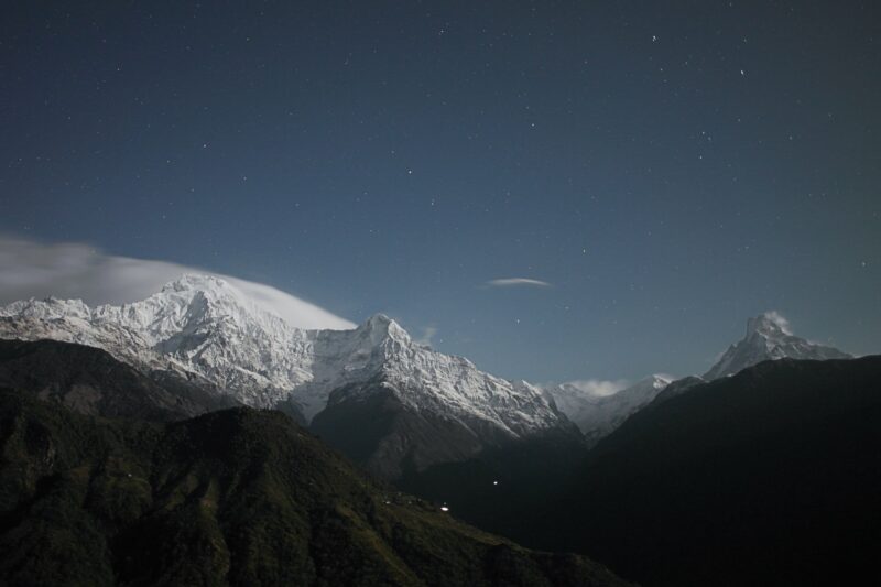 Snow-capped mountains under a starry night sky, with a dark valley in the foreground.