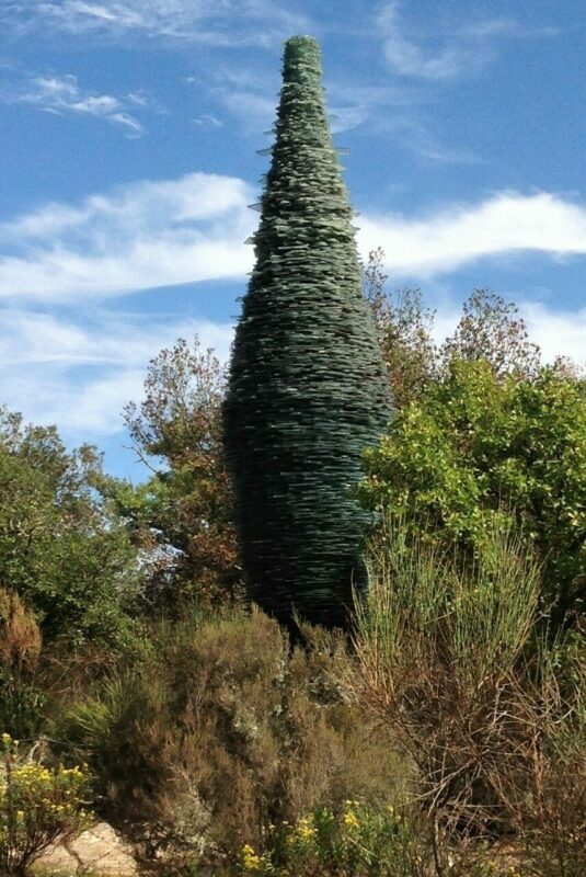 Tall, conical sculpture made of stacked glass pieces, surrounded by greenery and blue sky, at Chianti sculpture park.