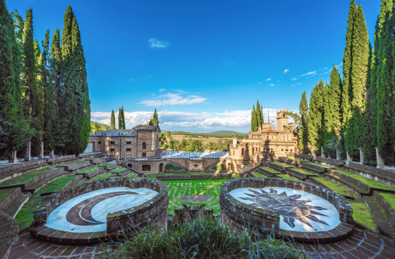 Panoramic view of Scarzuola, featuring stone structures, cypress trees, and symbolic designs on the lawn under a blue sky.