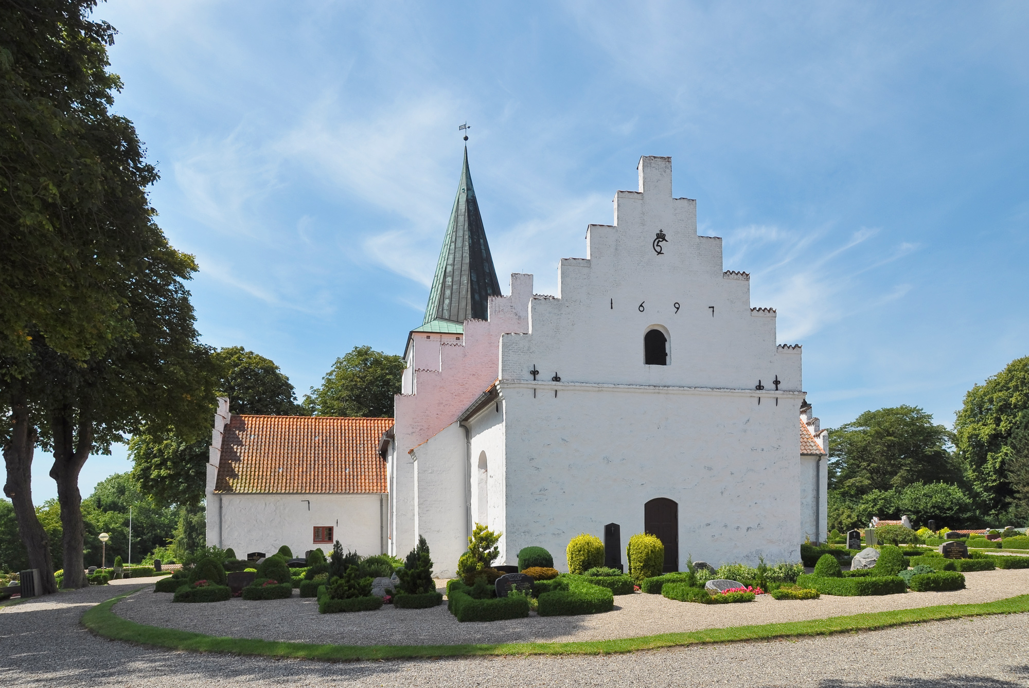 White church with a distinctive gabled roof and a green spire, surrounded by neatly trimmed hedges and trees.