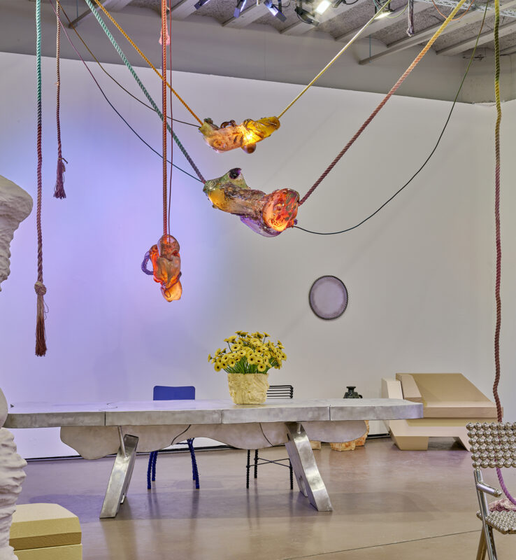 Art installation featuring a table with a sculptural base, illuminated hanging elements, and a vase of sunflowers.