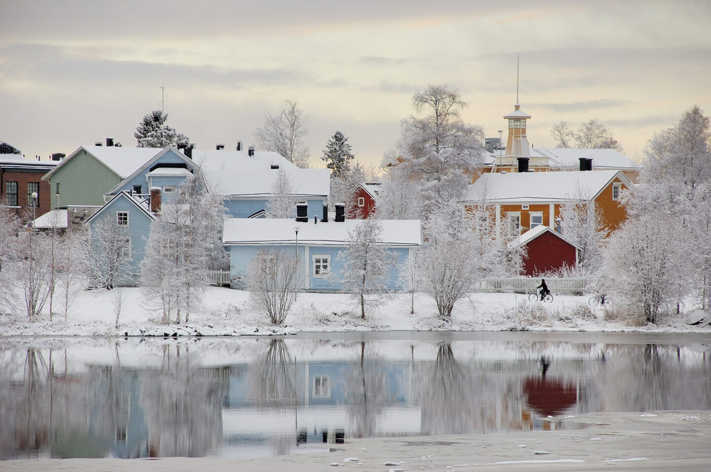 Colorful houses surrounded by snow and trees, reflecting in a calm river, depicting a snowy winter scene in Oulu.