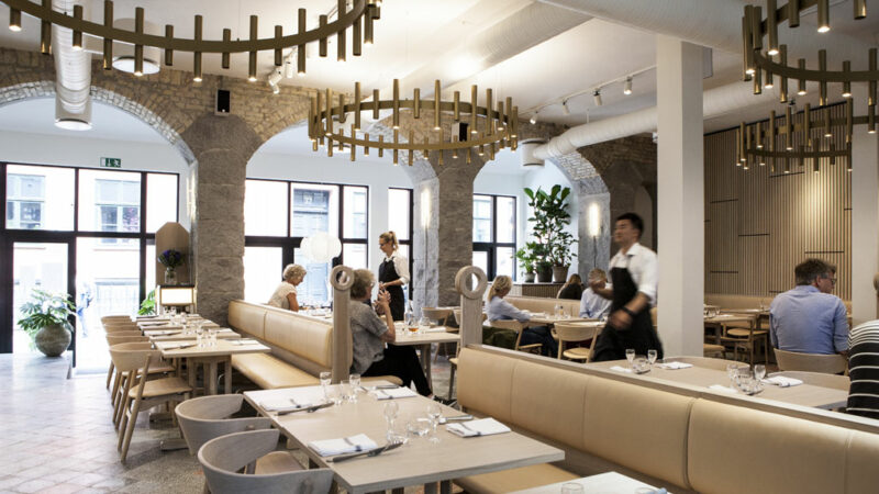 Modern restaurant interior with wooden decor, patrons dining, and staff serving in a bright, airy space.