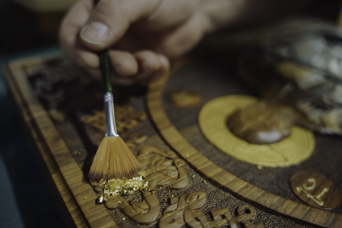 Hand applying gold leaf to a wooden carving, with intricate details visible on the surface.