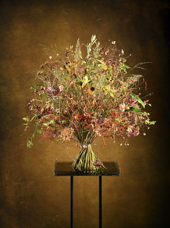 Colorful bouquet of dried flowers arranged in a glass vase on a black stand, set against a warm, textured background.