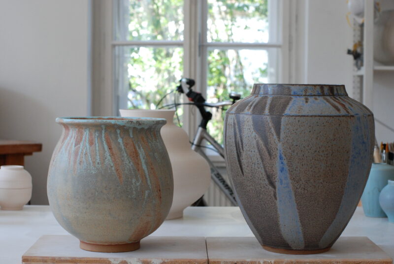 Two ceramic vases in a studio setting, one with a light blue glaze and drip patterns, the other with a textured brown finish.