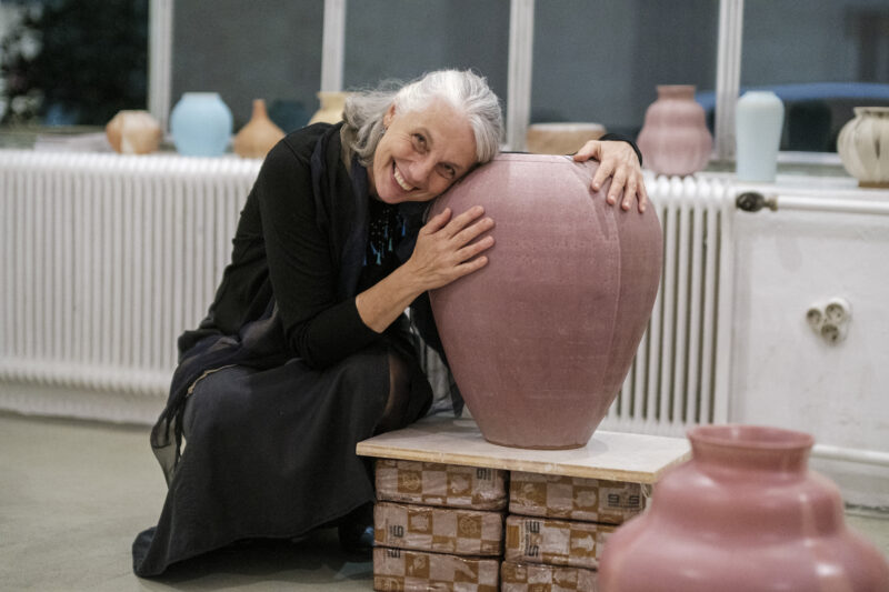 Woman with long gray hair embraces a large pink pottery vase, smiling in an art studio filled with ceramic pieces.