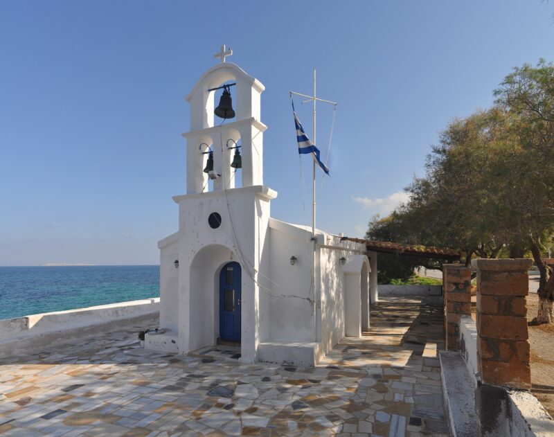 Unique white church with blue doors and a bell tower, overlooking the sea on Aegina Island, Greece.