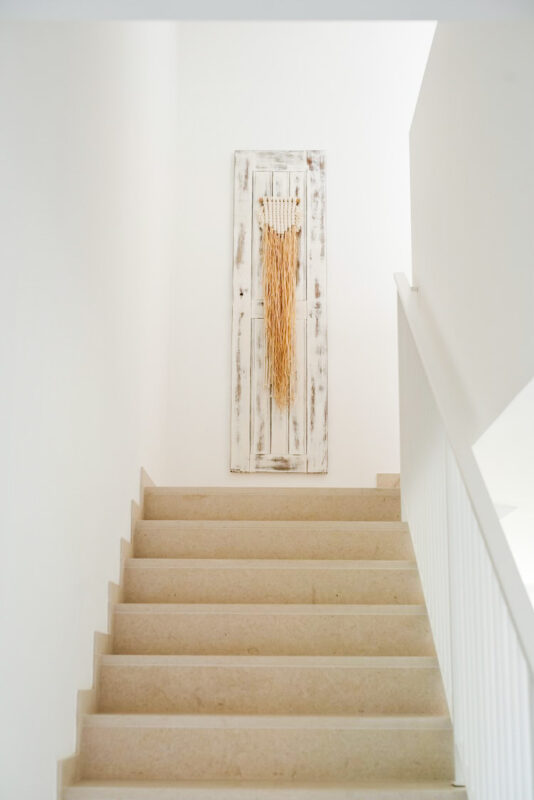 Staircase leading upwards with light-colored walls and a decorative wall hanging featuring long, natural fibers.