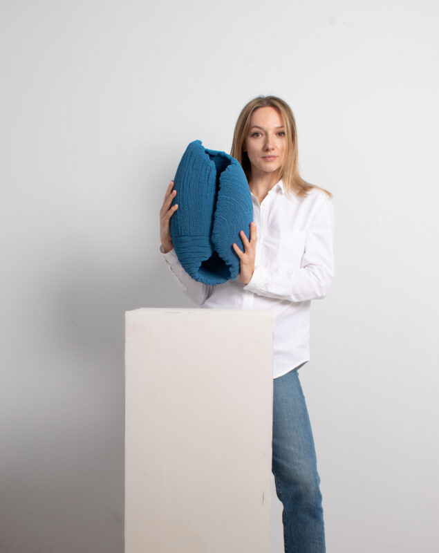 Woman in a white shirt holds a large blue textile object while standing next to a white pedestal.