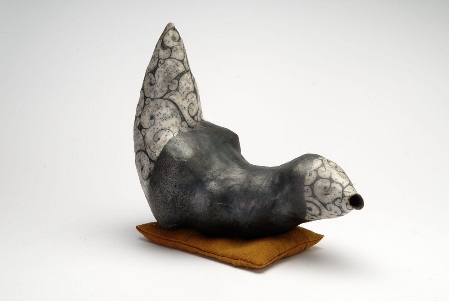 Ceramic sculpture with a stylized animal form, featuring a gray body and intricate black patterns, resting on a brown cush...