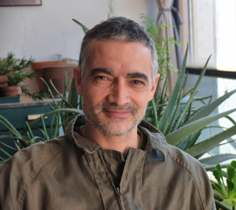 Man with short gray hair and a beard, smiling, seated in front of green plants indoors.