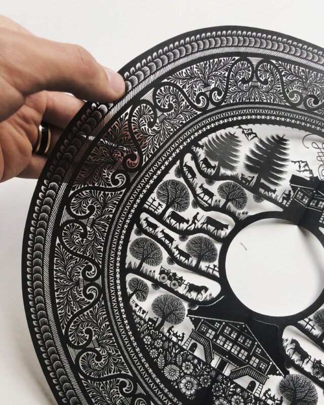 Intricate black paper cut art featuring rural scenes, held by a hand, with detailed patterns and a central circular cutout.