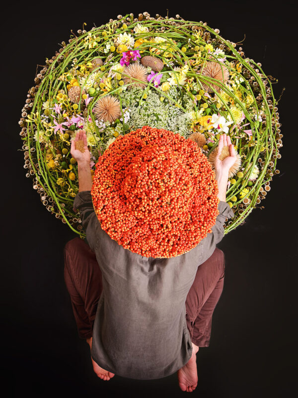 Person kneeling on a black surface, surrounded by a circular arrangement of colorful flowers and greenery, wearing a large...