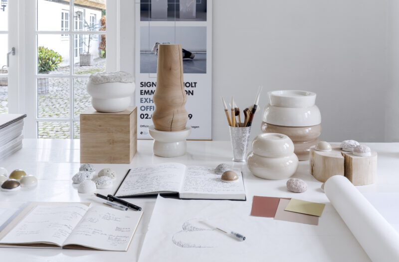 Artistic workspace featuring sketches, ceramic pieces, and materials on a table, with a window view in the background.