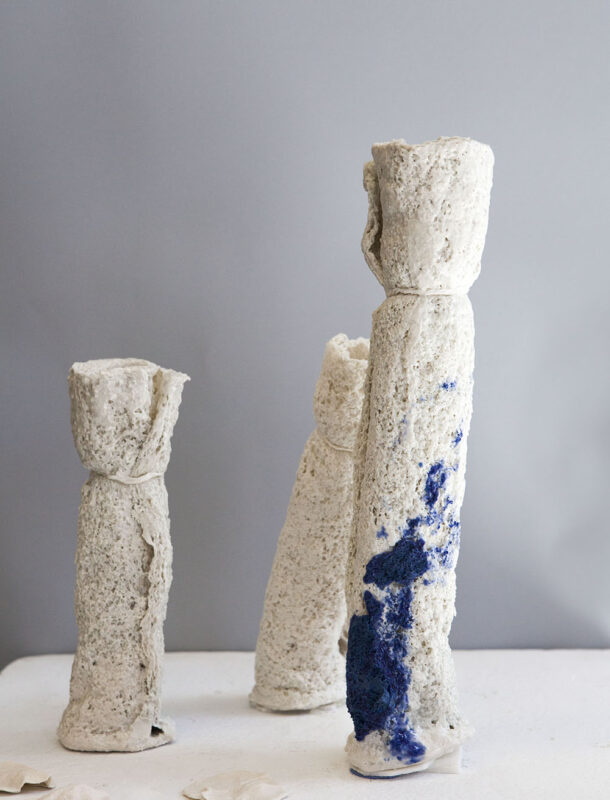 Three textured, cylindrical sculptures in neutral tones, one with blue accents, set against a gray background.