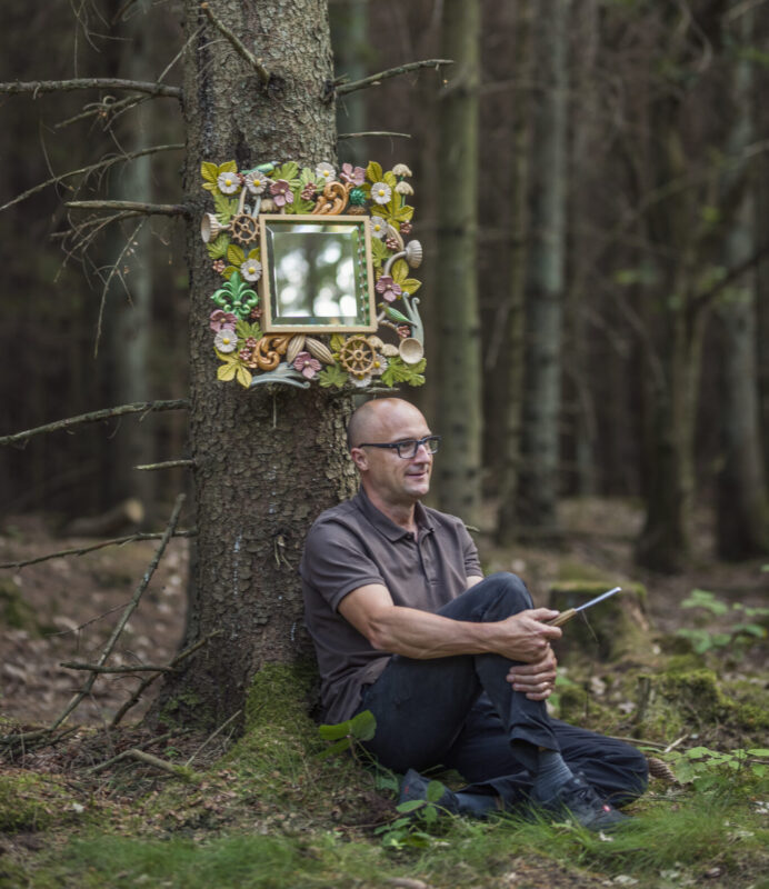 Man sitting against a tree in a forest, with a decorative mirror mounted on the tree behind him.