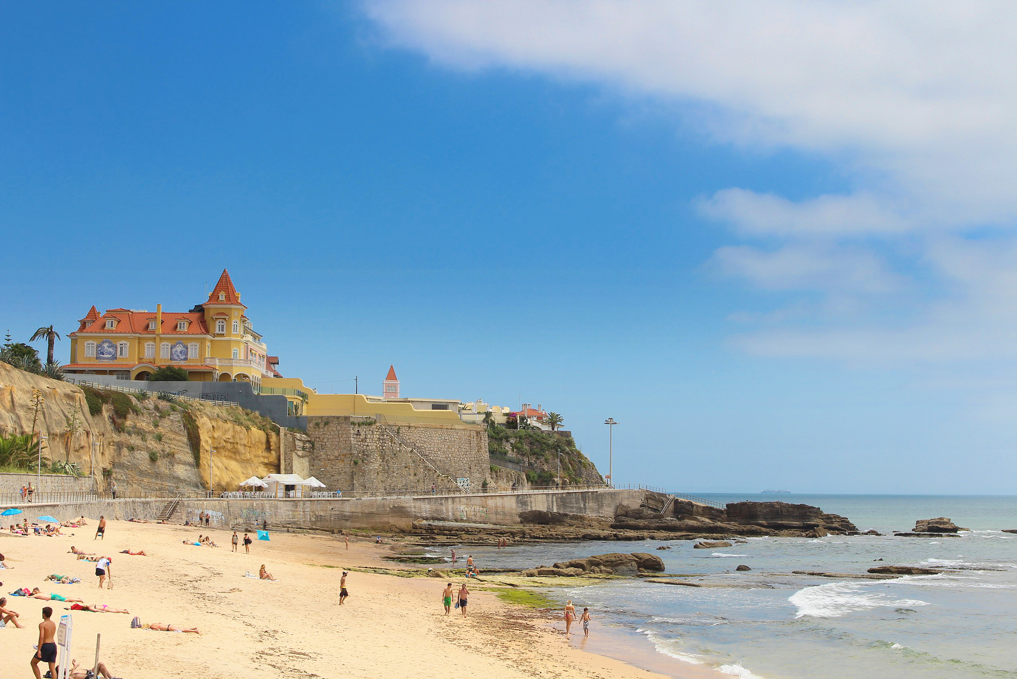 Beach near Lisbon featuring a sandy shore, rocky outcrops, and a colorful building on a cliff under a blue sky.