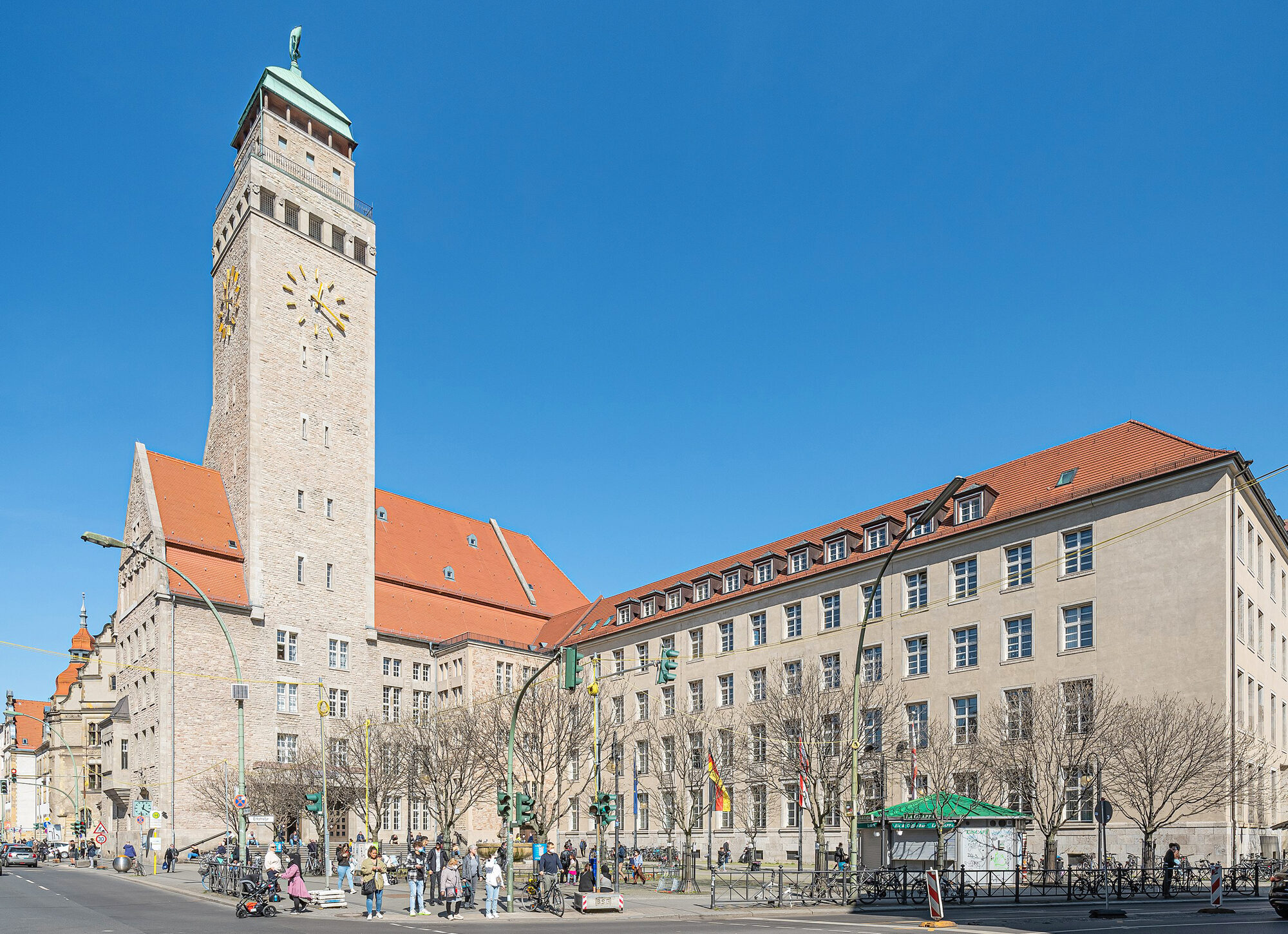 Neukölln Rathaus in Berlin, featuring a tall clock tower and modern buildings, with people and trees in the foreground.