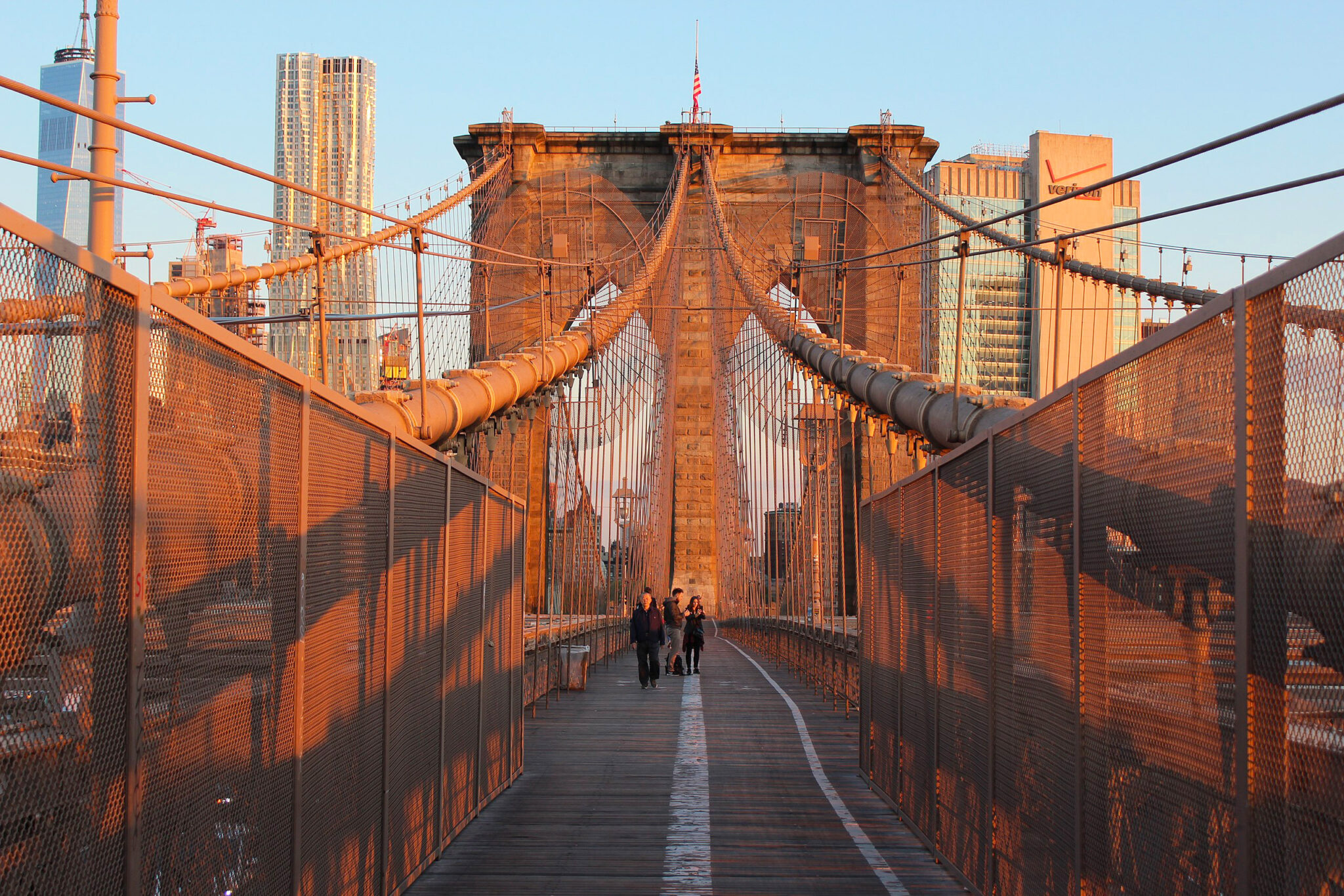 Brooklyn Bridge upper deck walkway at sunset, featuring pedestrians and city skyline in warm tones.