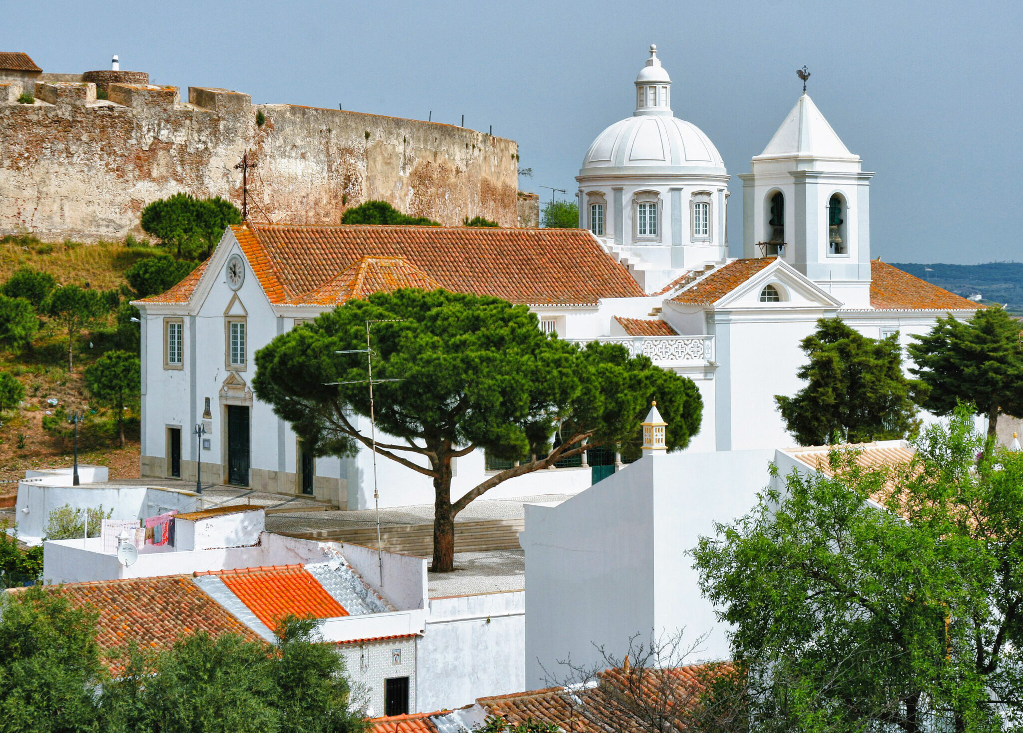 White buildings with red roofs and a dome, surrounded by greenery, set against a castle wall in Castro Marim, Algarve.
