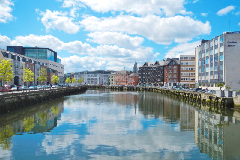 Panoramic view of the River Lee in Cork, Ireland, with reflections of buildings and a blue sky with clouds.
