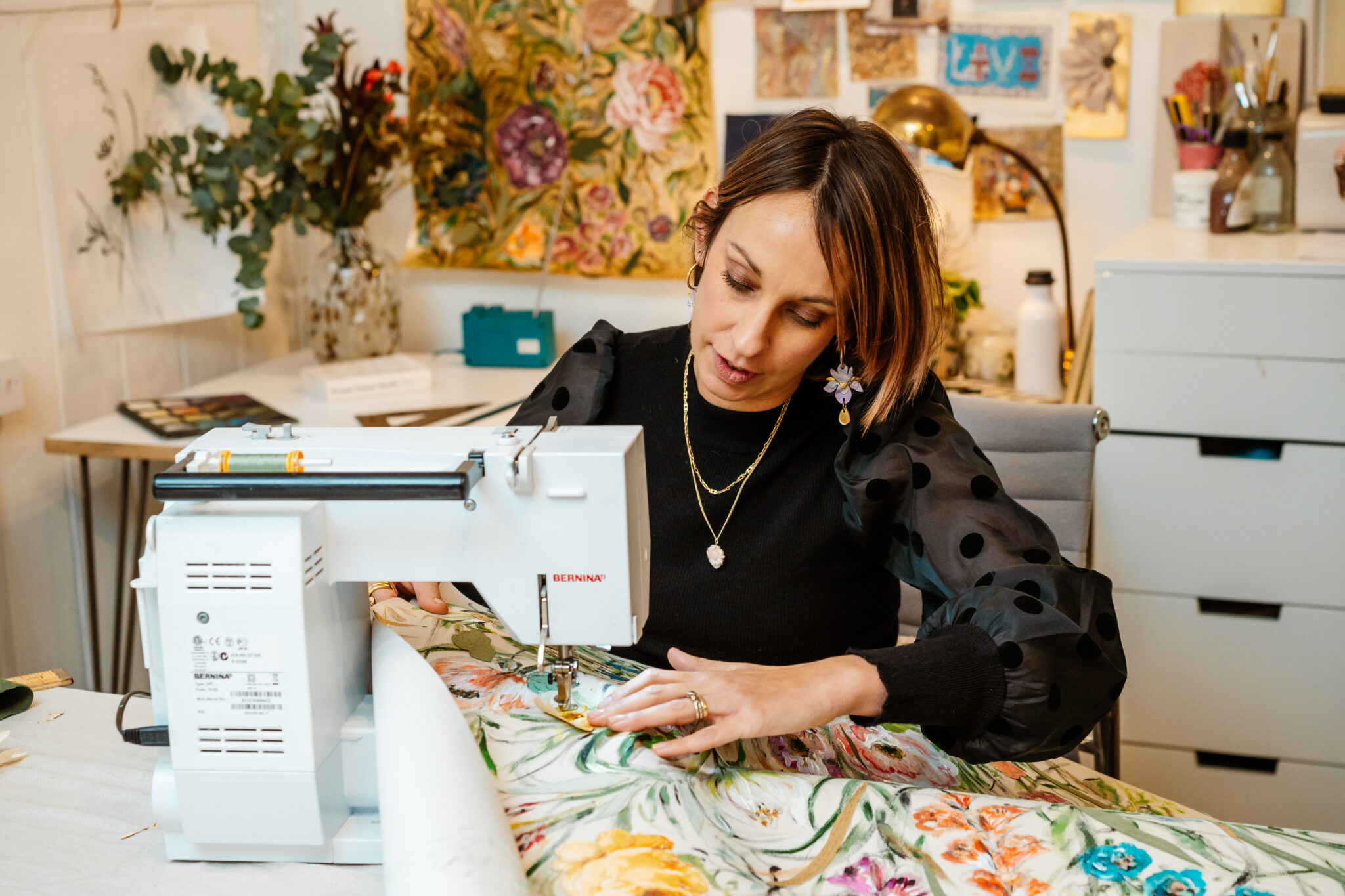 Woman sewing on a machine, surrounded by floral fabric and artistic decor in a creative workspace.