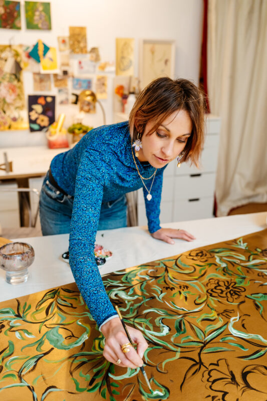 Artist Claire Coles painting on a large, ornate fabric in a bright studio, surrounded by colorful artwork.