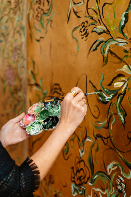 Artist's hand holding a palette with paint, applying details to a floral mural on a brown wall.