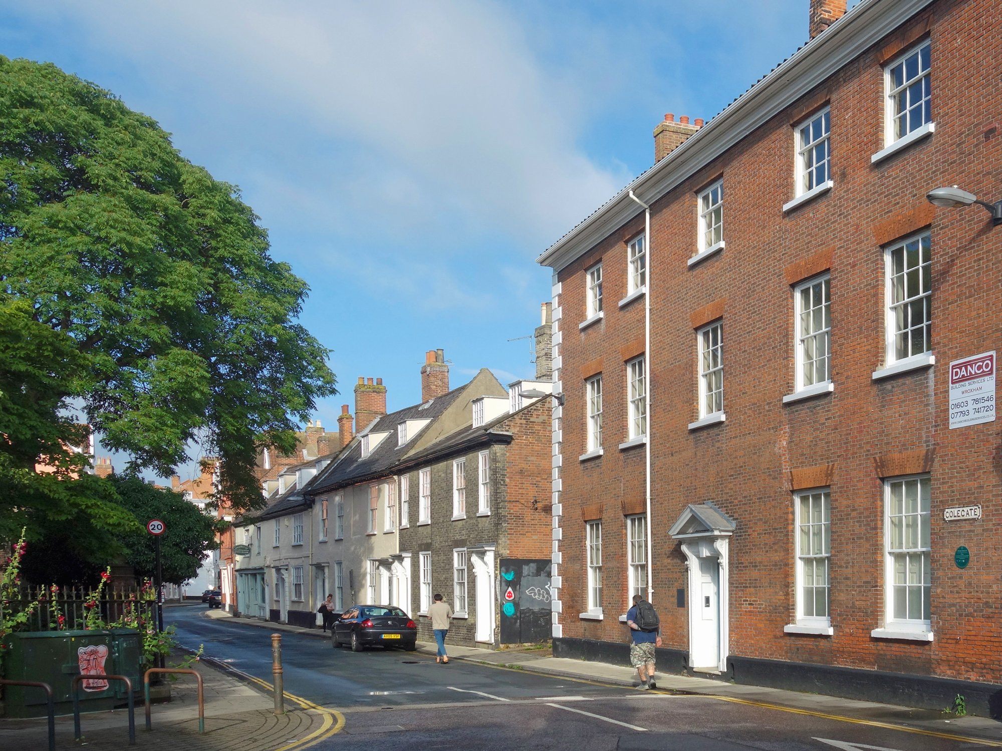 Historic street view in Norwich featuring brick buildings, a tree, and parked cars under a blue sky.