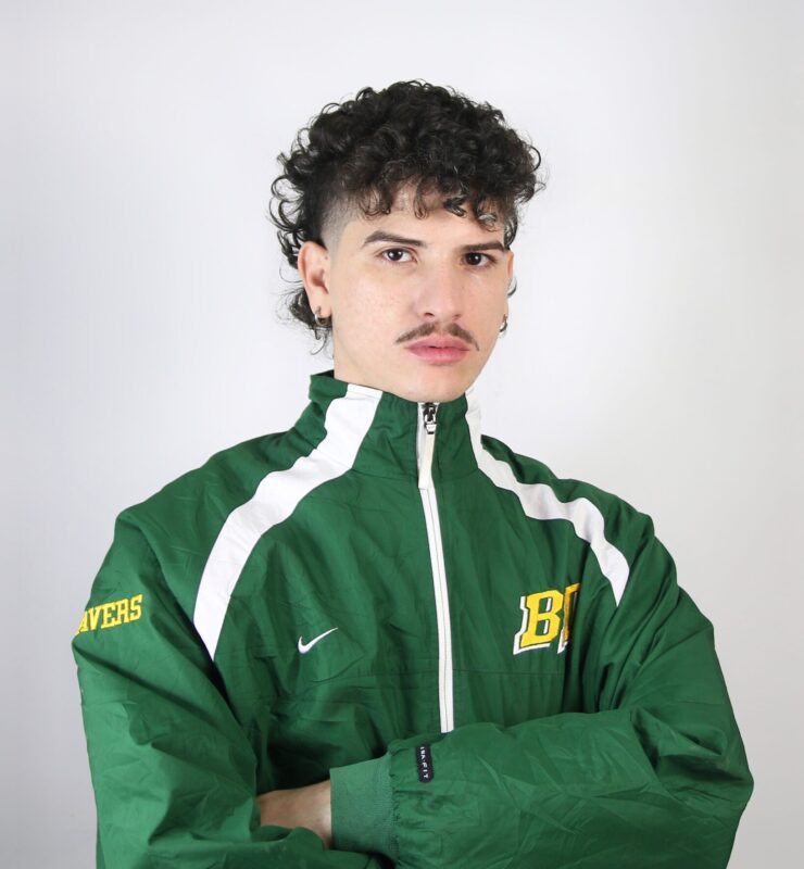 Young man with curly hair and a mustache, wearing a green athletic jacket with white accents and a logo.