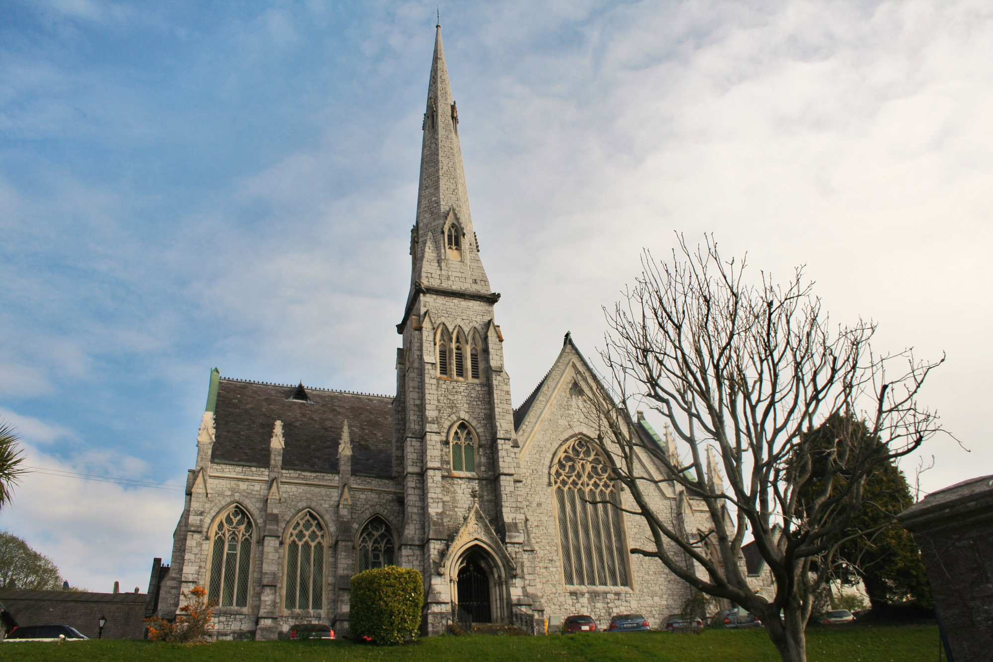 Gothic-style Cork Trinity Presbyterian Church with a tall spire, surrounded by trees and a cloudy sky.