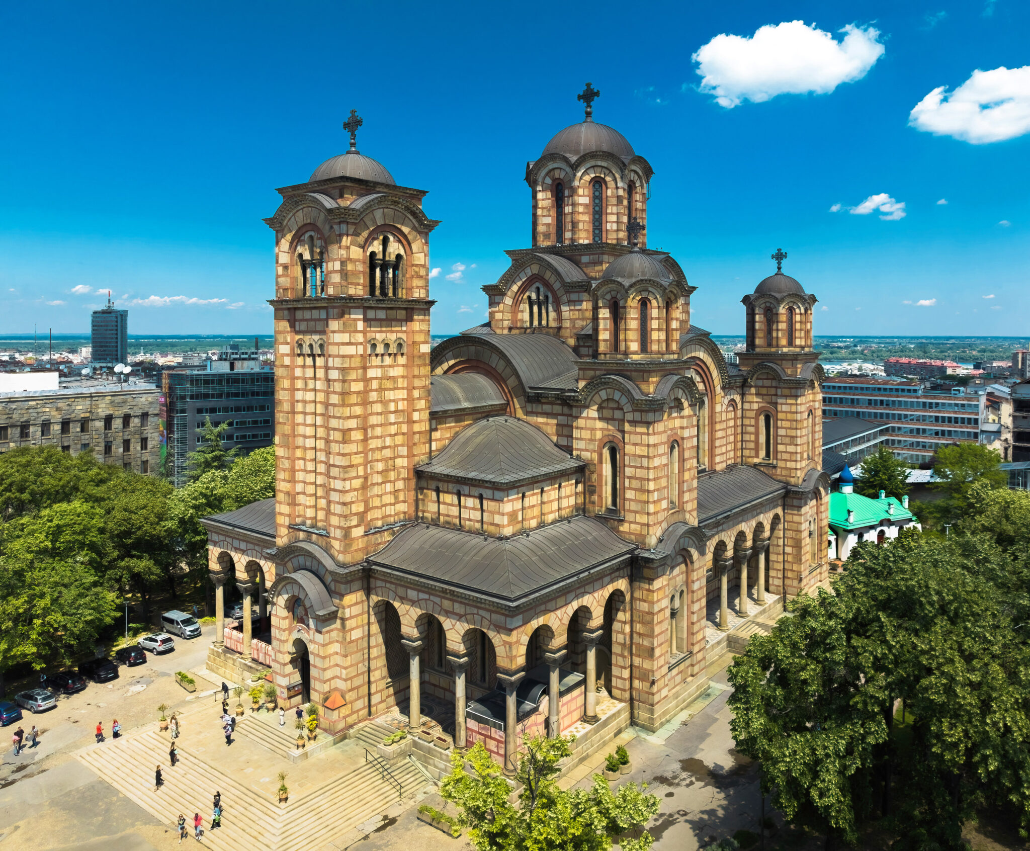 St. Mark's Church in Belgrade, featuring distinctive architecture and surrounded by trees and visitors on a sunny day.