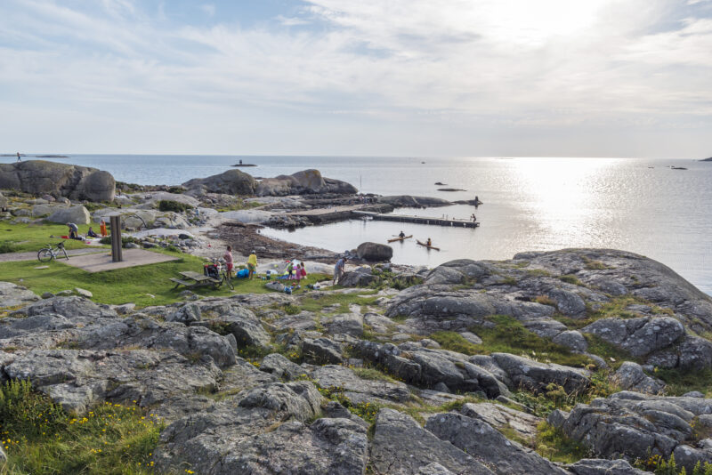 Rocky beach at Hästen on Hönö island, with people enjoying the sea breeze and kayakers in the water.