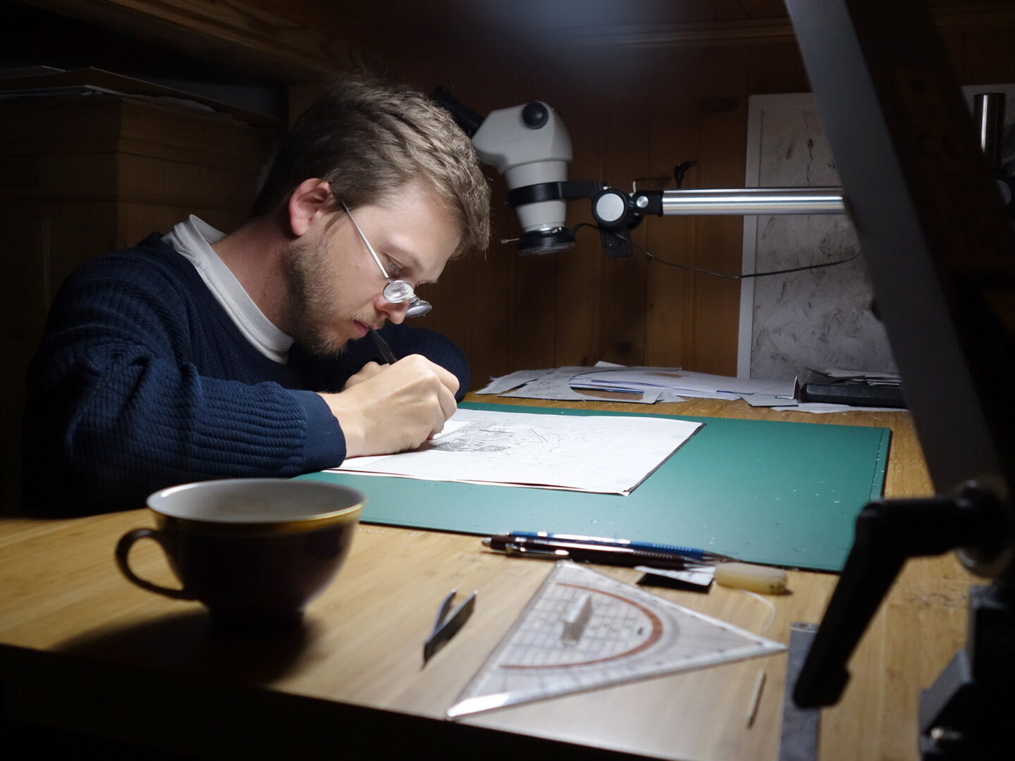 A young man with glasses sketches on paper at a wooden desk, with tools and a cup nearby, under focused lighting.