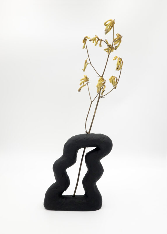 Black abstract sculpture with a wavy design, holding a dried yellow flower branch against a white background.