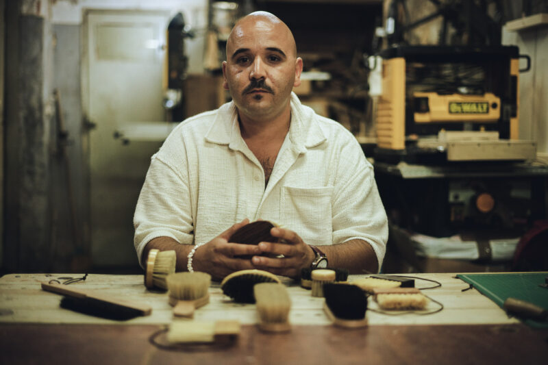 Man with a mustache seated at a table surrounded by various brushes and tools in a workshop setting.