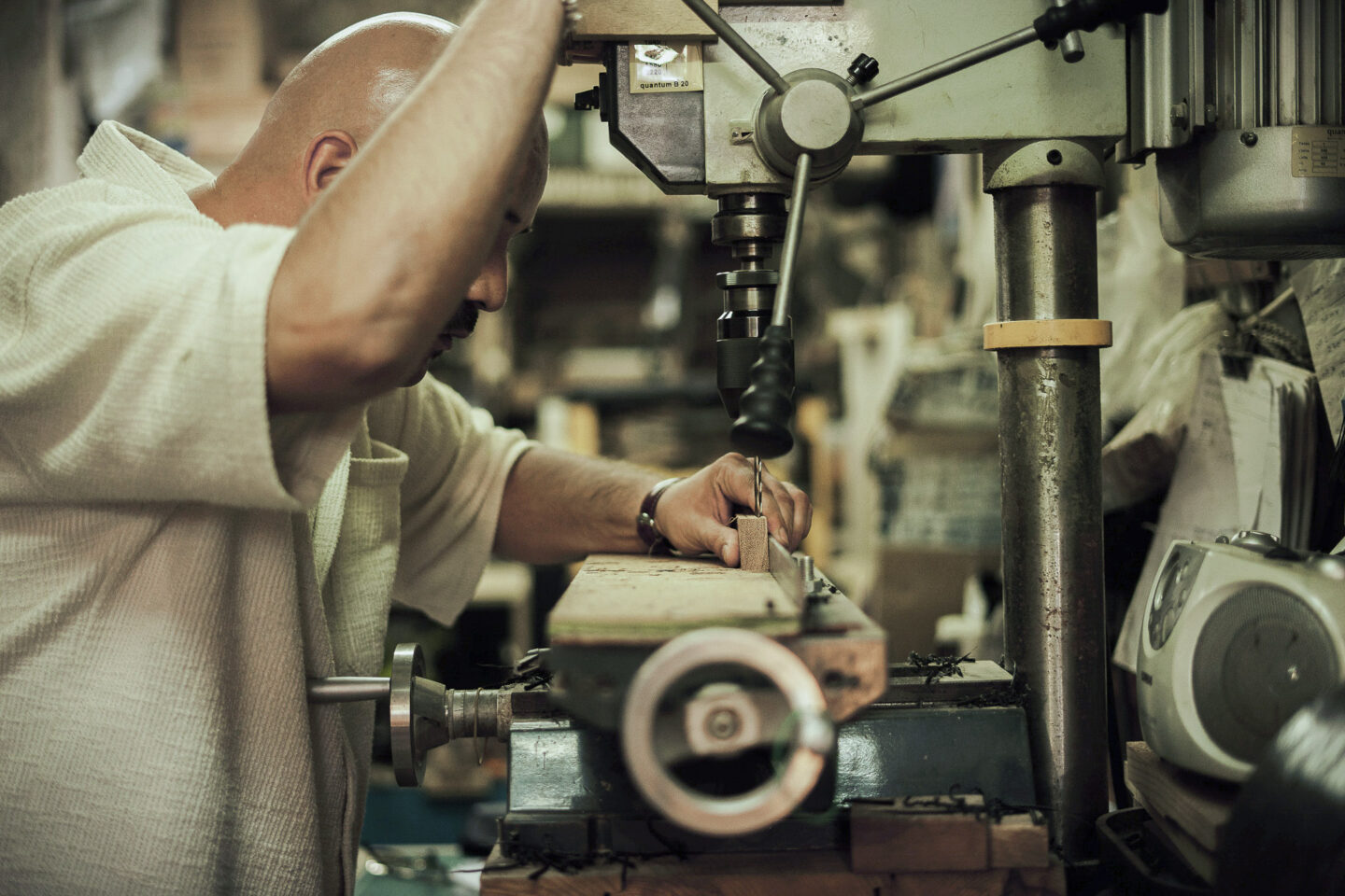Craftsman using a drill press to shape wood in a workshop filled with tools and materials.