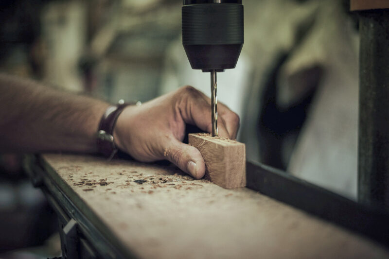 Hand holding a wooden block while drilling into it with a power drill in a workshop setting.