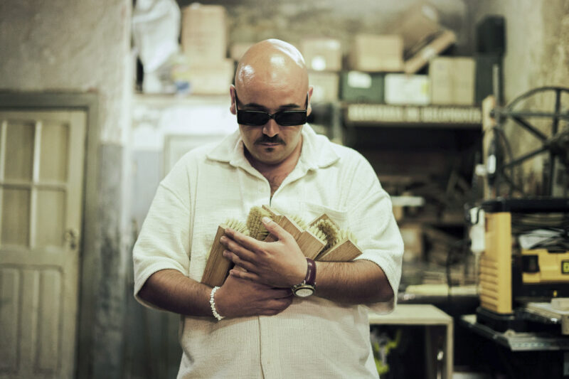 Bald man in sunglasses holds wooden blocks close, standing in a cluttered workshop.