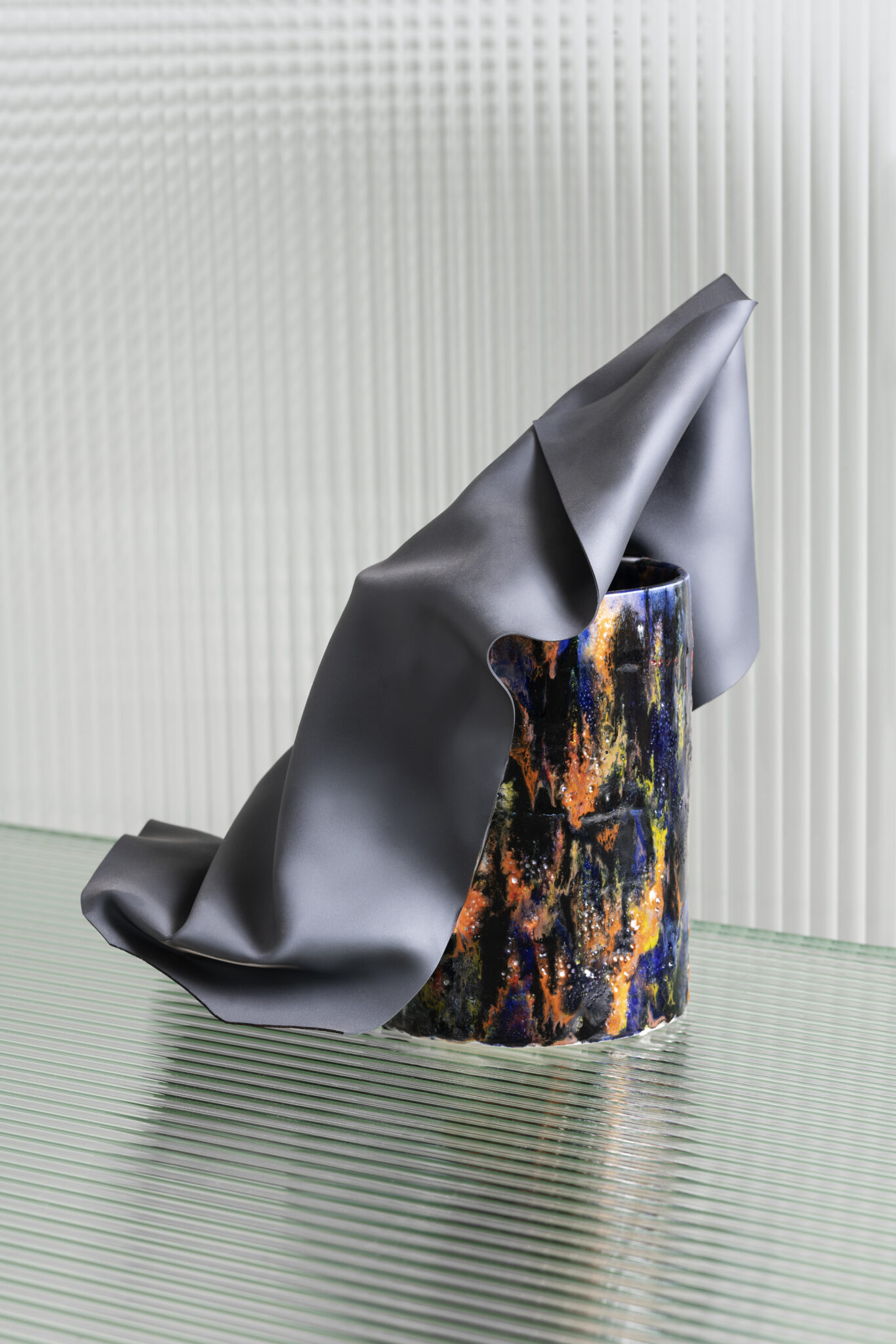 Minimalist vase with colorful glazing draped in gray fabric, set against a textured glass background.