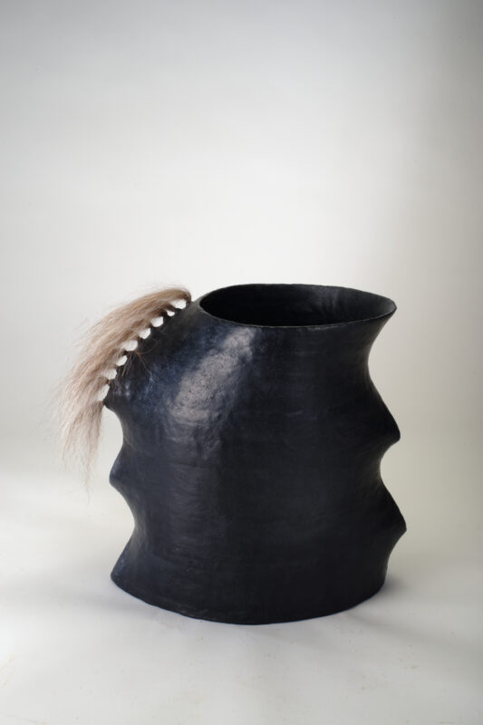 Black sculptural vessel with a wavy form and a tuft of hair attached to the rim.