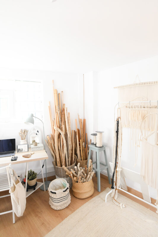 Bright, minimalist workspace featuring a desk with a laptop, baskets of sticks, and macramé supplies against white walls.