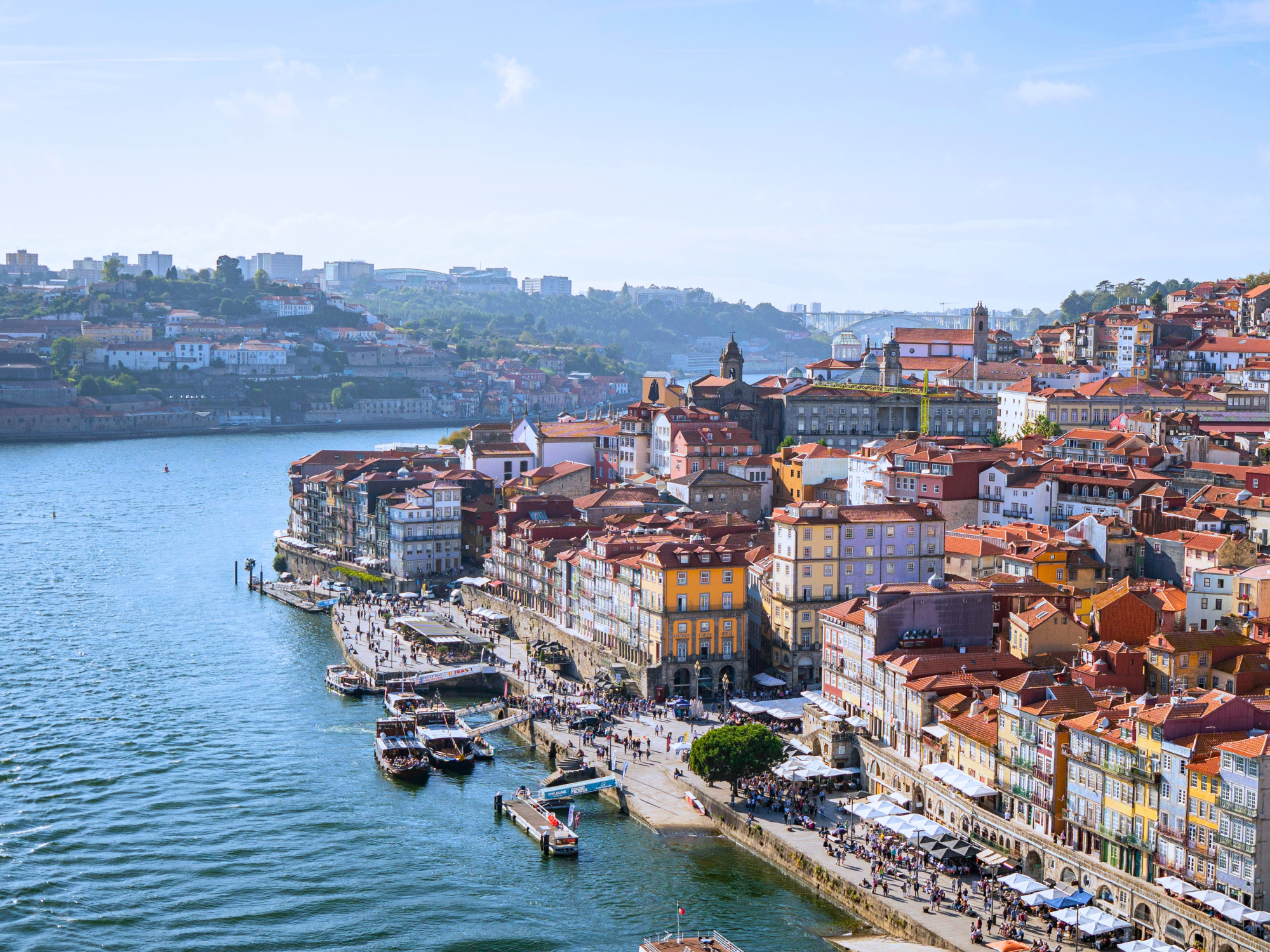 Colorful buildings line the riverbank in Porto, Portugal, with boats on the water and hills in the background.