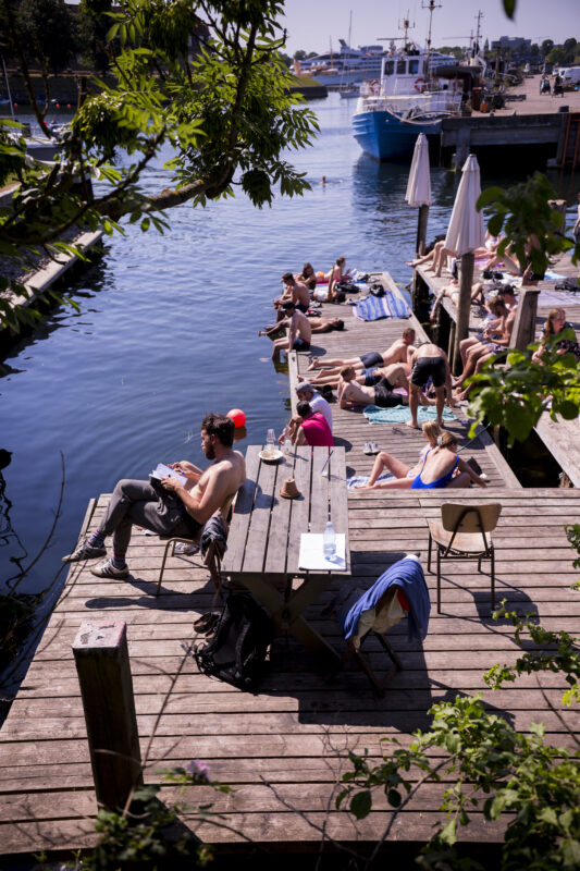 People sunbathing on wooden docks by a calm harbour, with boats and umbrellas in the background.