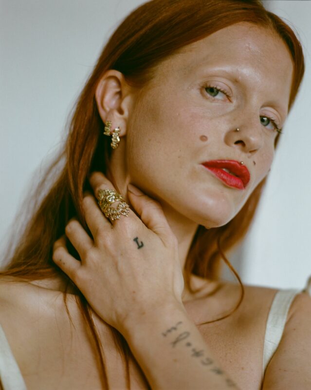 Close-up of a person with red hair, wearing rings and earrings, showcasing jewelry by Cameron.