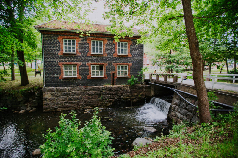 Historic stone building with red brick accents beside a flowing stream, surrounded by lush green trees in Fiskars, Finland.