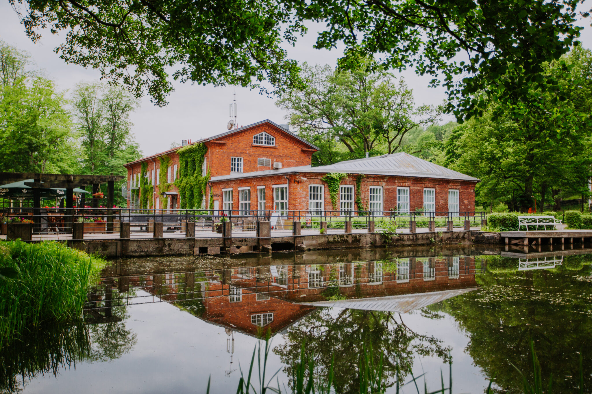 Red brick building with large windows reflecting in a calm pond, surrounded by lush greenery in Fiskars village.