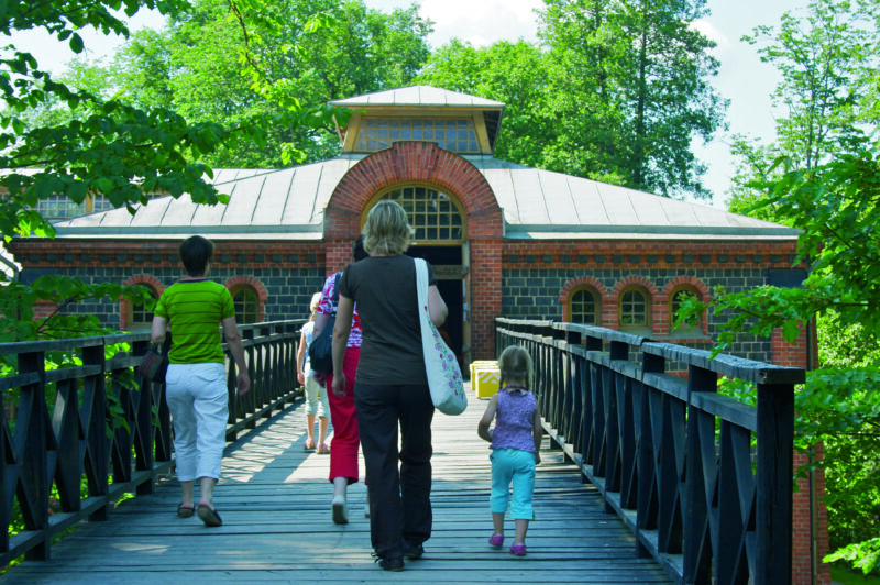 Four people walking on a wooden bridge towards a brick building surrounded by greenery on a sunny day in Fiskars.