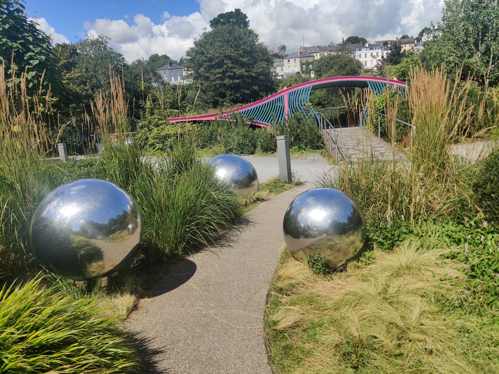 Reflective silver spheres on a pathway in Fitzgeralds Park, with a colorful bridge and greenery in the background.